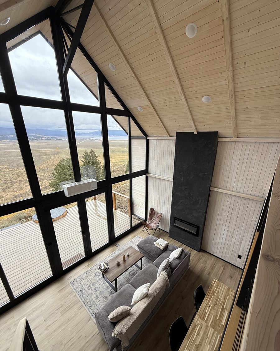 View from the loft over the living area, tall windows and black fireplace inside an Aries 116 cabin in Island Park, ID.
