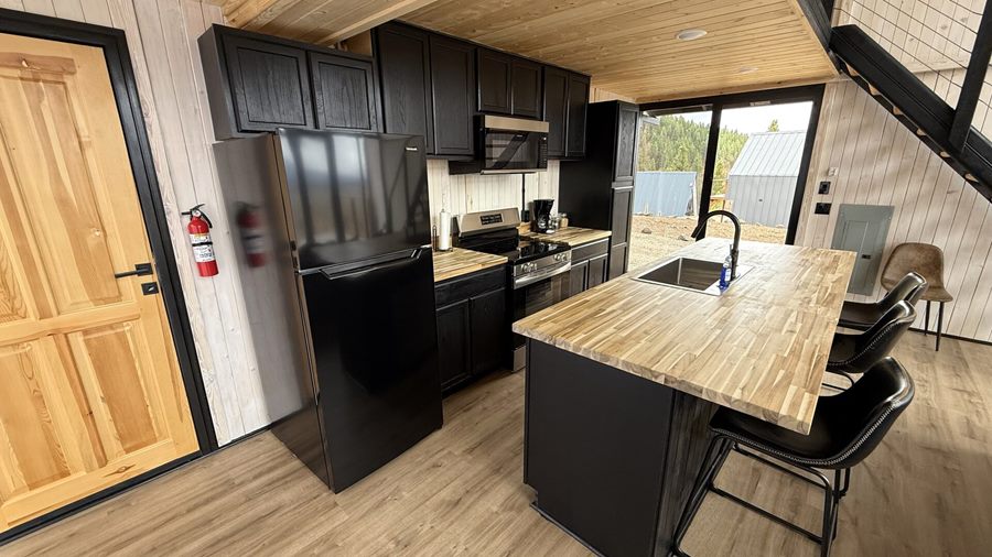 Wide view of a kitchen with black cabinetry, wood island and bar stools inside an Aries 116 cabin in Island Park, ID.
