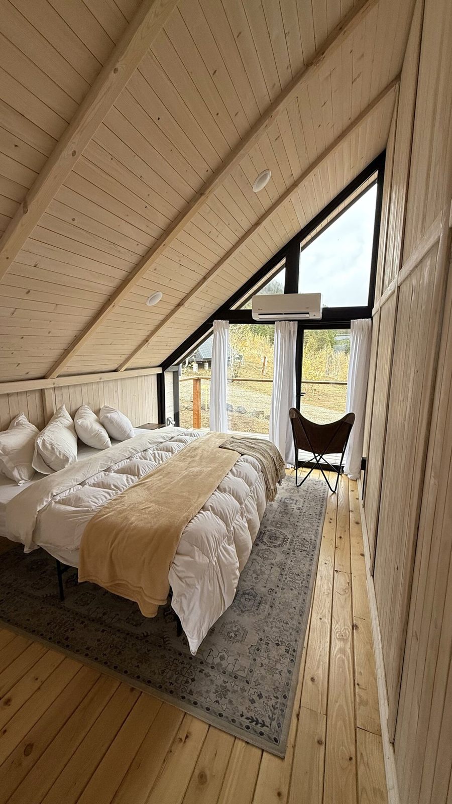 Vertical view of a furnished loft bedroom with bed, rug and tall triangular window inside an Aries 116 cabin in Island Park, ID.