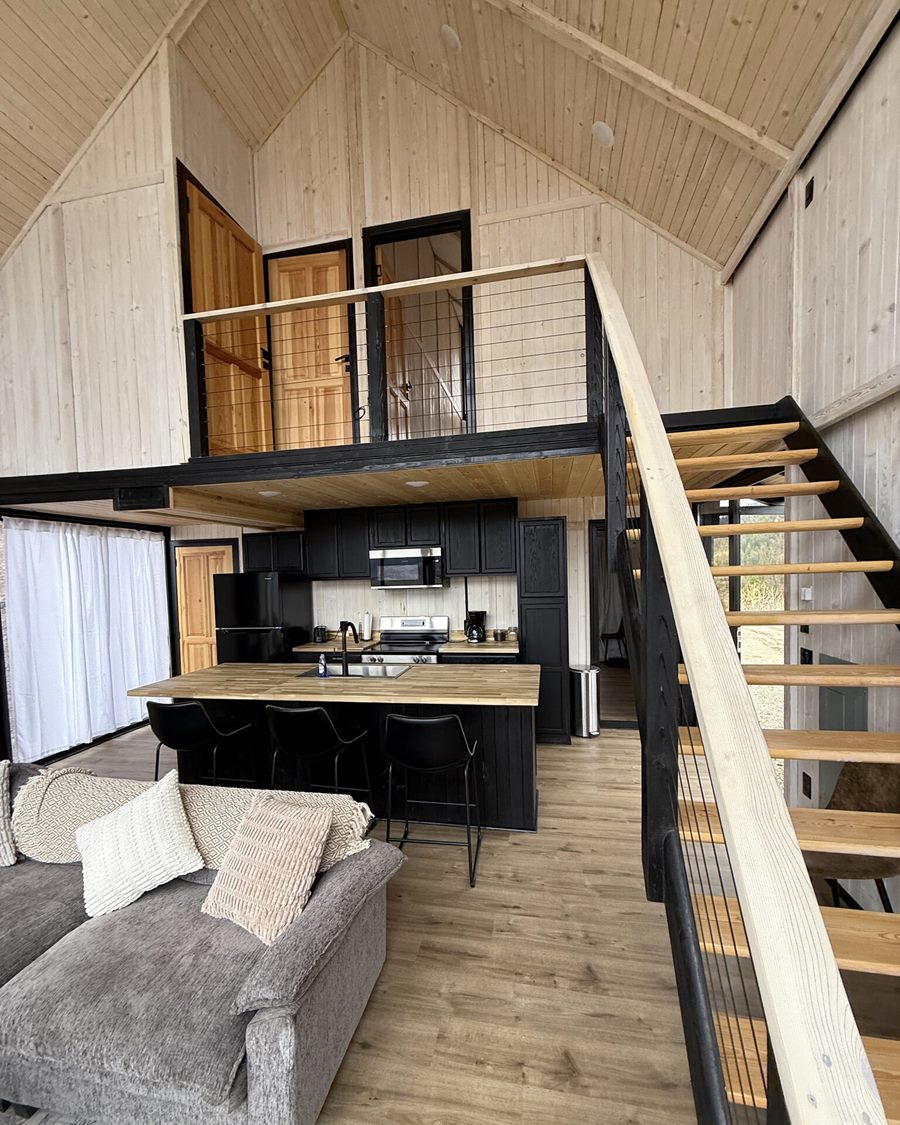Open-plan living area with kitchen, staircase and loft railing inside an Aries 116 cabin in Island Park, ID.