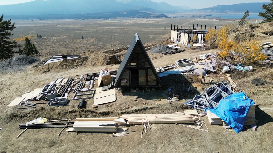 Cabin components and building materials distributed around a Stella 53 unit on the sloped Island Park, Idaho site.
