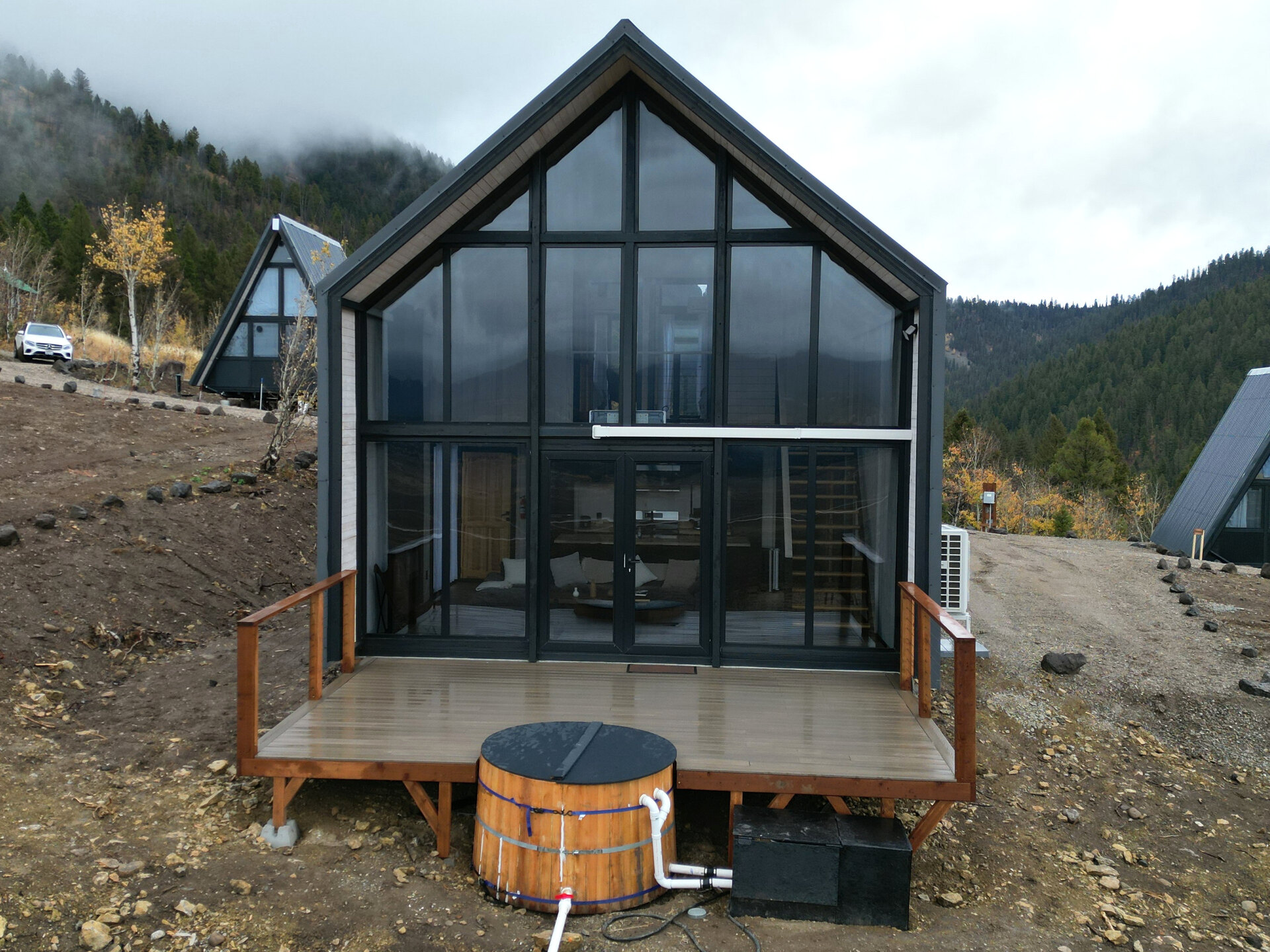 Front aerial view of an Aries 116 cabin with floor-to-ceiling glazing, a deck and a round hot tub in Island Park, Idaho.